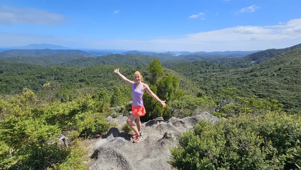 Sandra Tanner on a rocky lookout above native bush on Great Barrier Island, arms out, the sea behind her